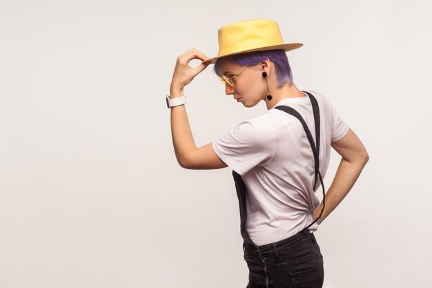 Portrait de vue latérale d'une élégante fille hipster élégante aux cheveux violets et en salopette tenant un chapeau sur la tête, regardant l'espace vide pour votre publicité. isolé sur fond blanc, tourné en studio