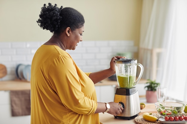 Portrait de vue latérale d'une femme noire en surpoids à l'aide d'un mélangeur tout en faisant un smoothie sain à la maison