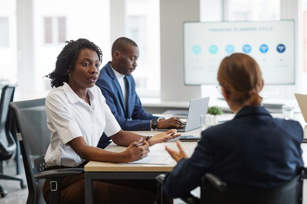 Portrait de vue latérale d'une jeune femme afro-américaine parlant à un collègue utilisant un fauteuil roulant lors d'une réunion d'affaires au bureau