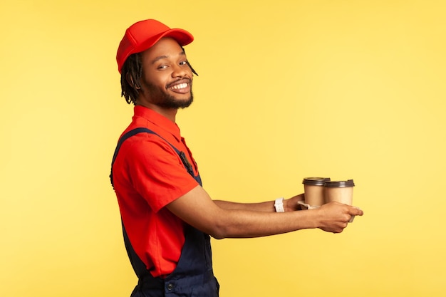 Portrait de vue latérale d'un messager positif souriant portant un T-shirt rouge et un uniforme bleu, donnant du café à emporter au client, bon de livraison. Studio intérieur tourné isolé sur fond jaune.