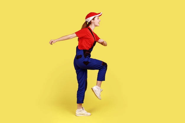 Portrait de vue latérale d'une ouvrière marchant, allant au travail, exprimant des émotions positives et du bonheur, portant une salopette et une casquette rouge. Studio intérieur tourné isolé sur fond jaune.