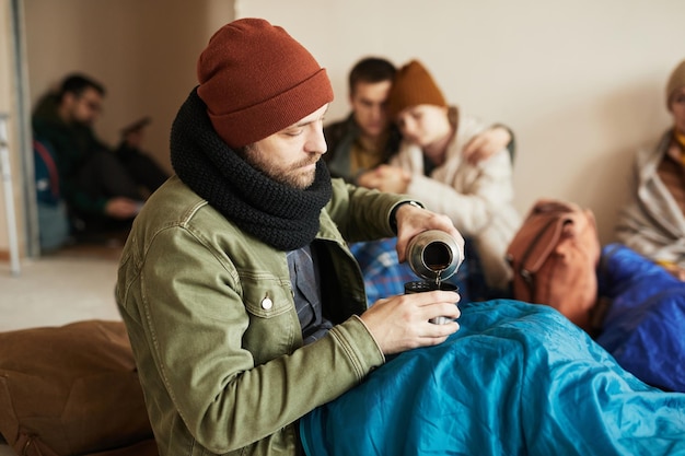 Portrait de vue latérale d'un réfugié masculin barbu versant du thé à partir d'un thermos tout en se cachant dans un abri couvert