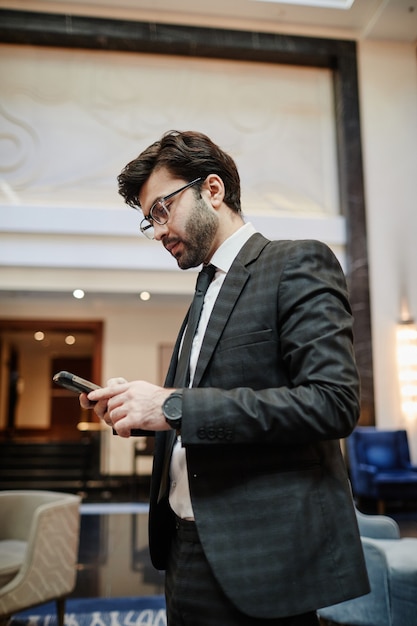 Portrait de vue latérale verticale d'un homme d'affaires prospère tenant un smartphone tout en se tenant dans le hall de l'hôtel