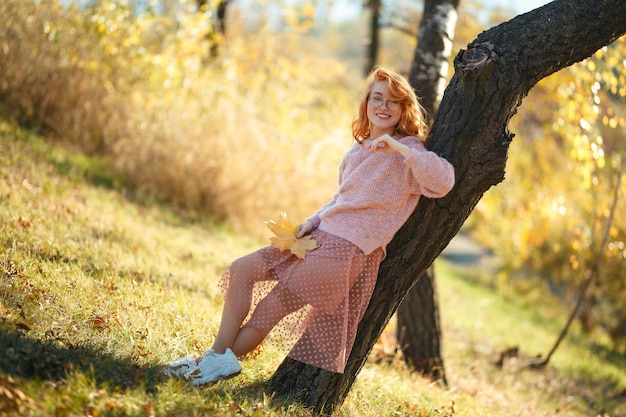 Portraits d'une charmante fille aux cheveux roux avec un joli visage. Fille posant dans le parc automne dans un pull et une jupe de couleur corail. Dans les mains d'une jeune fille une feuille jaune