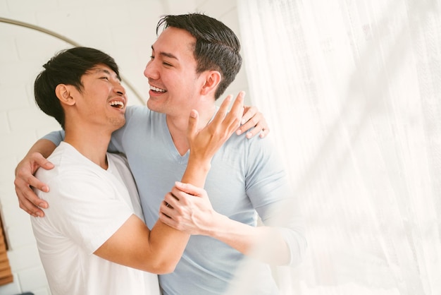 Portraits d'un couple gai asiatique du millénaire heureux étreignant et embrassant souriant et riant dans une fenêtre à rideau blanc dans le salon de la maison multirelationnelle LGBT Un concept de couple gay