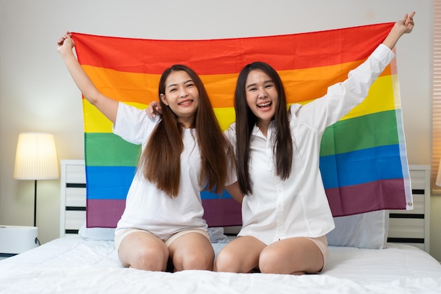 Photo portraits d'un couple de lesbiennes asiatiques heureux assis avec un drapeau arc-en-ciel assis à côté du lit