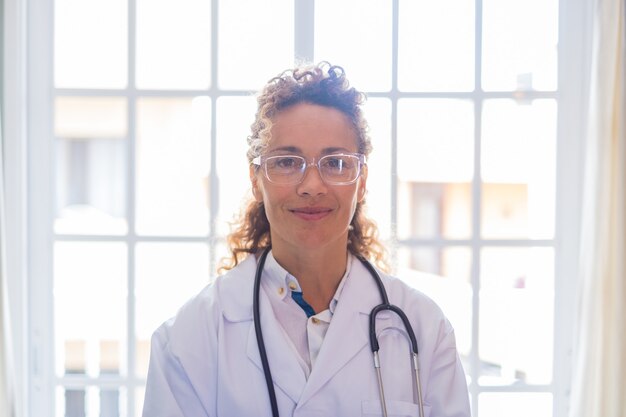 Portriat de jeune femme médecin en uniforme et stéthoscope debout contre la fenêtre à la clinique. Médecin de première ligne ou travailleur de la santé confiant dans des lunettes à l'hôpital.