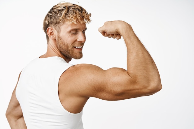 Portrit de bel athlète masculin vue arrière fléchissant les biceps et souriant montrant un bras fort après l'entraînement dans la salle de gym fond blanc