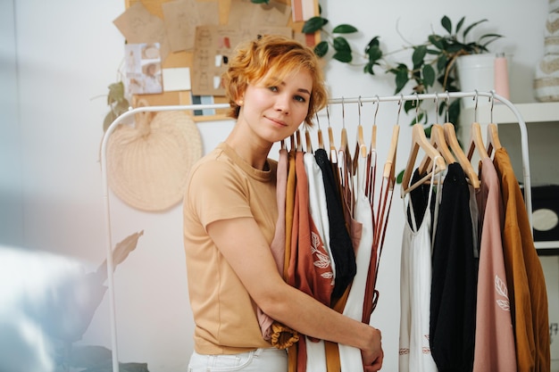 Posant une jeune couturière étreignant ses robes sur le rack Heureuse de son travail Elle a les cheveux courts teints au gingembre