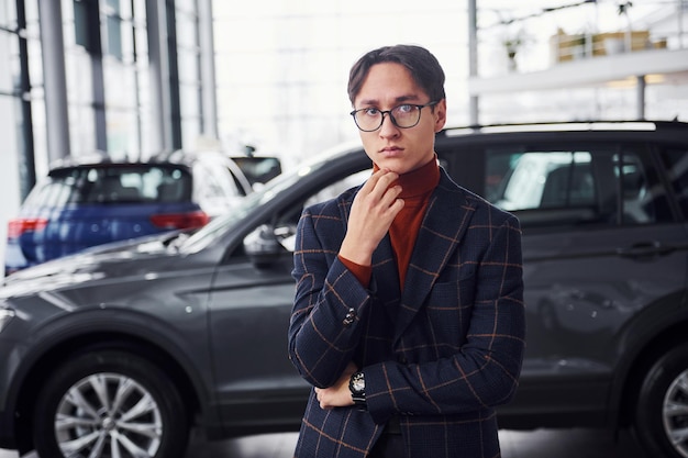 Posant pour la caméra. Jeune homme d'affaires en costume de luxe et vêtements formels est à l'intérieur près de la voiture.