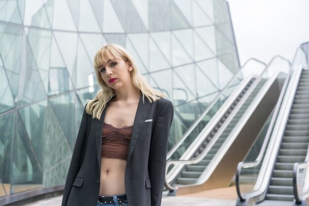 Pose à la mode d'une jeune blonde dans des bâtiments en verre de la ville