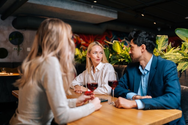 Positif deux femmes blondes et bel homme barbu en costume et assis à table dans un restaurant parlant s'amusant à célébrer l'anniversaire