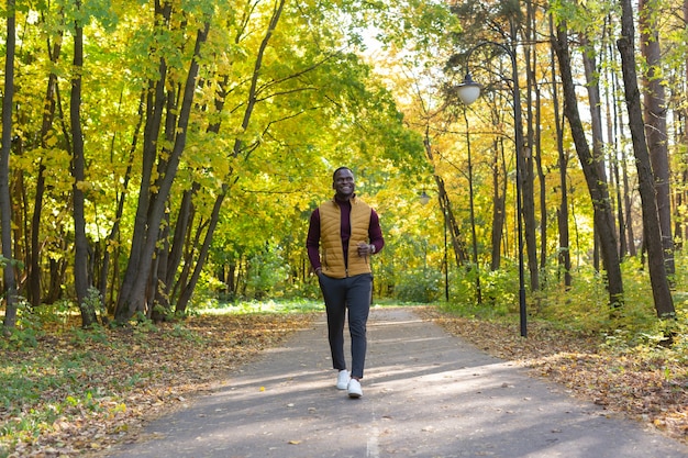 Positif jeune homme afro-américain dans des vêtements élégants se promène dans le parc de la fin de l'été sous le soleil