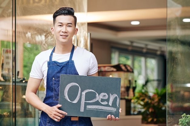 Positif jeune homme asiatique debout à l'entrée du café avec panneau ouvert