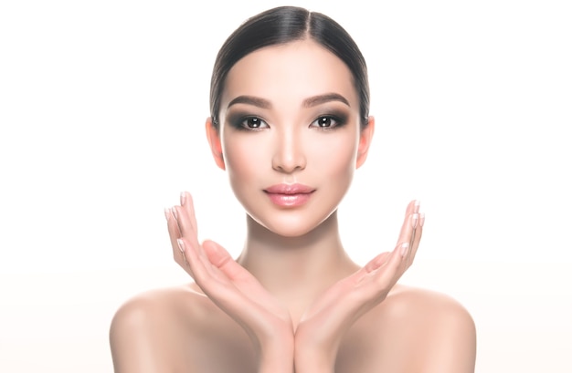 Position symétrique des mains devant le visage de la belle jeune femme asiatique aux épaules nues Maquillage délicat avec des yeux charbonneux et des lèvres roses Traitement du visage cosmétologie beauté et spa