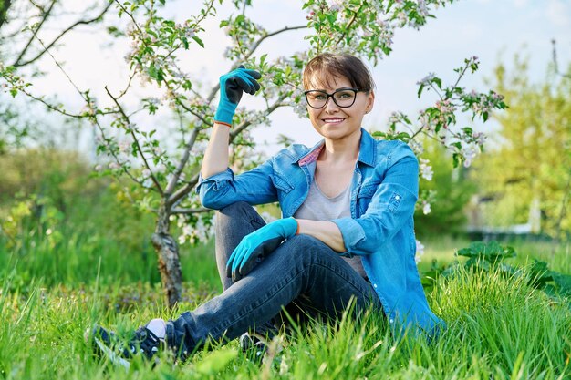 Positive belle jardinière mature assise sur l'herbe avec un pommier en fleur femme d'âge moyen souriante en gants de jardinage se reposant après le travail de printemps saisonnier personnes de 40 ans printemps