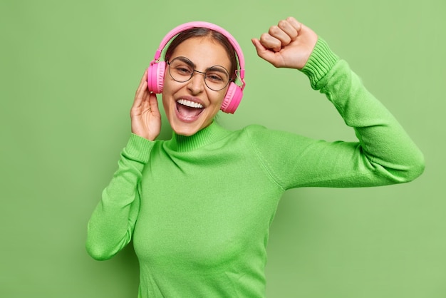 Positive belle jeune femme aux cheveux noirs se déplace sans soucis attrape chaque morceau de chanson utilise un casque stéréo porte des lunettes rondes col roulé décontracté isolé sur fond vert vif