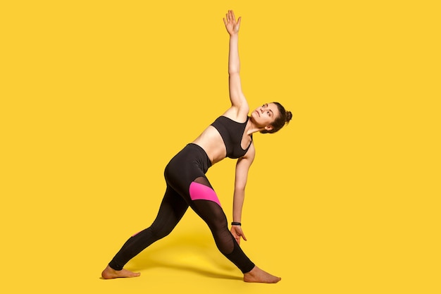 Posture triangulaire. Femme athlétique mince avec chignon dans des vêtements de sport serrés pratiquant le yoga, faisant de l'exercice Trikonasana, pliant et étirant les muscles. prise de vue en studio sur toute la longueur, entraînements sportifs isolés