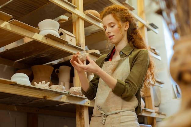 Pot en argile inhabituel. Belle femme aux cheveux longs intéressée en chemise verte tenant une petite création tout en recherchant des pièces intéressantes