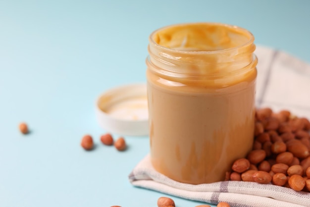 Un pot de beurre de cacahuète est posé sur une table bleue avec des cacahuètes dessus.