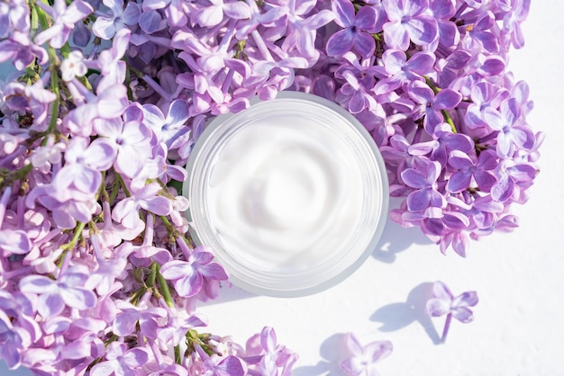 Pot blanc ouvert de crème légère à base de plantes pour femmes sur tableau blanc Belle branche de lilas violet Soin pour un visage propre et doux mains jambes et peau du corps Fleurs fraîches Place vide pour le texte