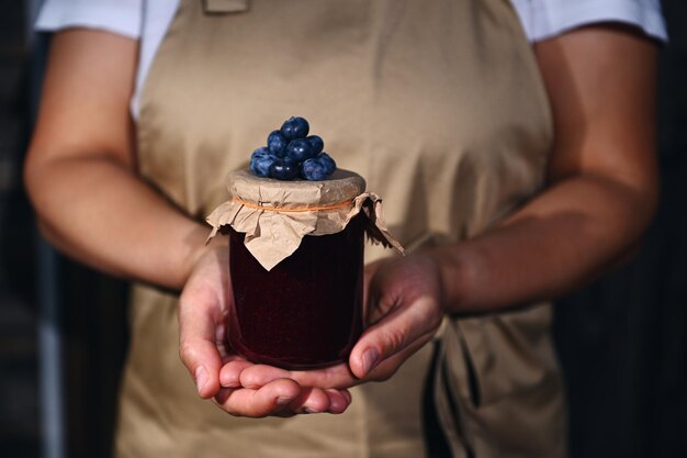 Un pot de confiture de bleuets avec des baies fraîches mûres sur un couvercle en papier kraft entre les mains d'une femme une femme au foyer confiseur