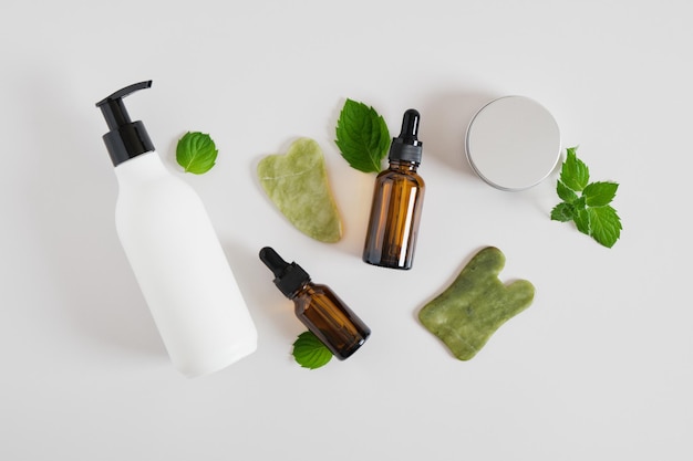 Pot de cosmétiques en aluminium, crème dans des flacons avec distributeur, sérum dans un flacon compte-gouttes et grattoir gua sha en pierre de jadéite, feuilles de menthe sur fond gris