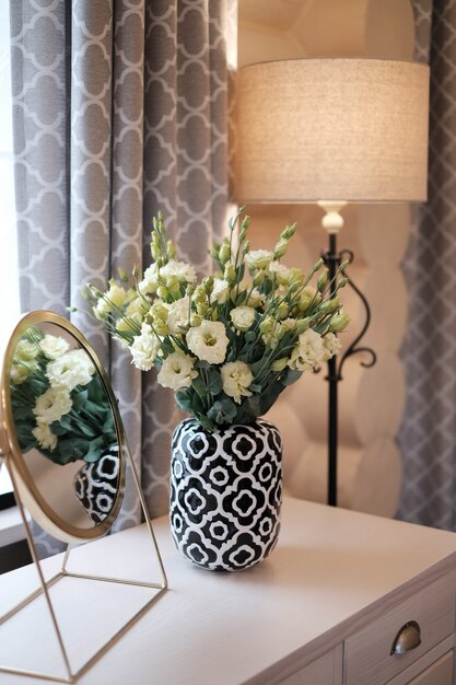 Pot de fleurs avec des fleurs sur une lampe de bureau blanche et un miroir près de la fenêtre dans un intérieur de maison moderne