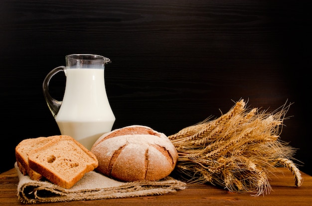 Pot de lait, pain de seigle rond, gerbe de blé et tranches de pain sur un sac, table en bois