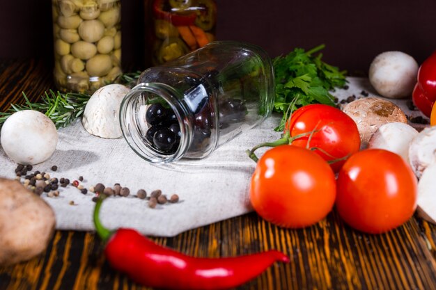 Pot ouvert avec des olives noires se trouve sur une serviette près des épices, tomates, champignons et autres légumes sur table en bois