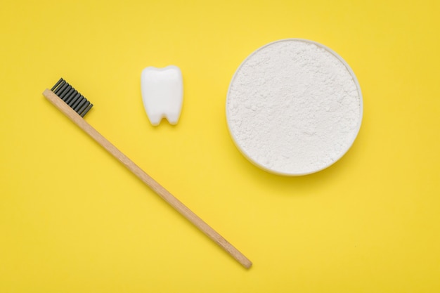 Un pot de poudre de nettoyage dentaire, une brosse à dents en bois et une figurine de dent sur une table jaune. La vue de dessus. Mise à plat.