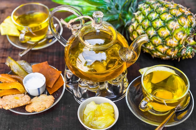 Pot de thé en verre avec du thé à l'ananas et des jam cups faits maison et des fruits cristallisés sur fond sombre