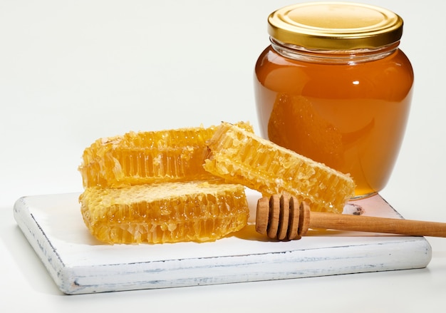 Pot transparent en verre avec du miel et des morceaux de nid d'abeilles en cire avec du miel sur une planche en bois blanche, fond blanc