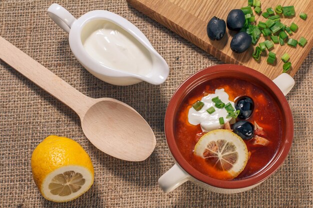 Potage salé avec viande, saucisses fumées, pommes de terre, tomates, concombre mariné mariné, citron, olives noires et crème sure dans un bol à soupe en céramique, planche à découper, cuillère en bois