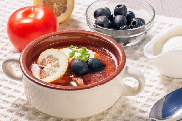 Potage salé avec de la viande, des saucisses fumées, des pommes de terre, des tomates, du concombre mariné mariné, du citron, des olives noires et de la crème sure dans un bol à soupe en céramique avec des ingrédients sur un torchon blanc