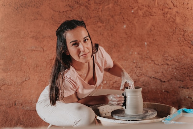 Une potière est dans son atelier en train de façonner le vase qu'elle fabrique avec un matériau appelé terre cuite