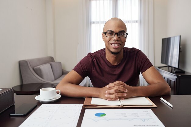 Potrait de beau jeune homme d'affaires noir souriant dans des verres assis au bureau à la maison et travaillant avec des documents financiers