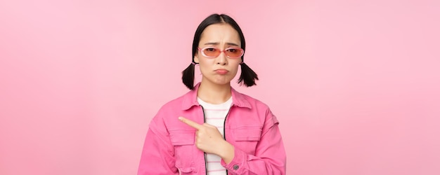 Potrait de jolie fille coréenne en lunettes de soleil pointant vers la gauche et regardant déçu bouder bouleversé debout sur fond rose