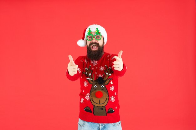 Pouce en l'air. bonne année. joyeux Noël. heureux homme barbu en chapeau de père noël. hipster brutal en pull de Noël tricoté en hiver. mec mature avec des lunettes de fête de vacances sur fond rouge.