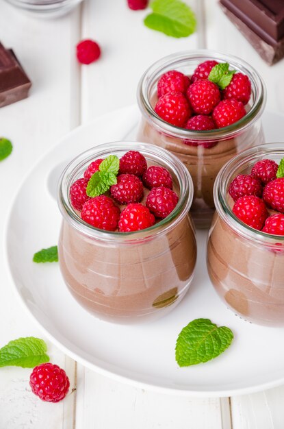 Pouding au chocolat (mousse) avec des framboises fraîches dans des bocaux en verre sur un bois blanc. Dessert pour la Saint-Valentin et la fête des femmes.