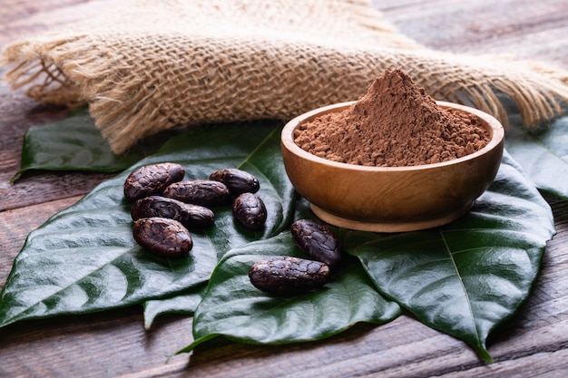 Poudre de cacao dans un bol en bois de fèves de cacao sèches entières et de feuilles fraîches sur une vieille table rustique