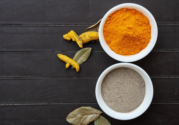 Poudre de curcuma et poudre de poivre noir dans des bols blancs, racines de curcuma et feuilles de laurier à côté des bols