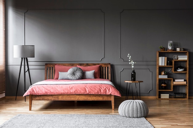 Pouf à côté d'un lit en bois avec des draps roses dans un intérieur de chambre gris avec étagère