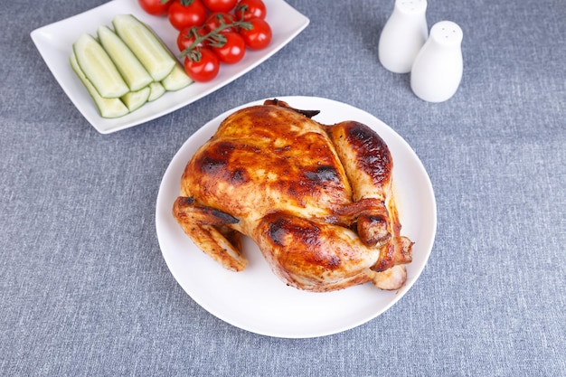 Photo poulet cuit entier sur une délicieuse croûte un plat traditionnel en arrière-plan se trouvent des concombres et des tomates cerises sur une assiette blanche gros plan mise au point sélective fond gris vue de dessus