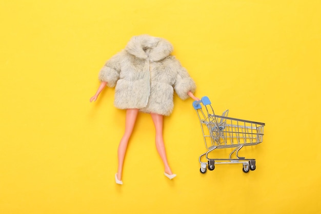 Poupée femme en manteau de fourrure et chaussures avec caddie sur fond jaune Mise en page de mode minimalisme Vue de dessus Mise à plat