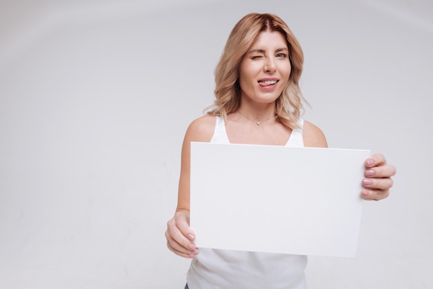 Pourquoi si sérieux. Remarquable femme séduisante artistique gaffant sur le plateau tout en faisant des grimaces et tenant un panneau blanc dans ses mains