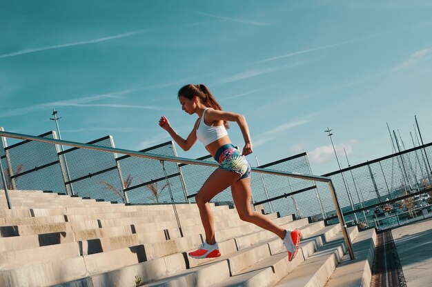 Pousser fort. Toute la longueur de la belle jeune femme en vêtements de sport courant tout en faisant de l'exercice à l'extérieur