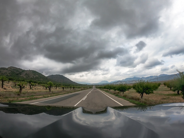 Pov conduisant une voiture noire sur une route goudronnée à double sens avec vue sur la campagne à Murcie avec des nuages bas noirs épars. Pour traverser une terre espagnole avec des montagnes d'un côté.