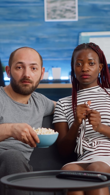 POV d'un couple interracial marié regardant un film d'horreur ensemble sur un canapé. Mari et femme multiethniques mangeant du pop-corn ayant peur en regardant la caméra sur le canapé du salon.