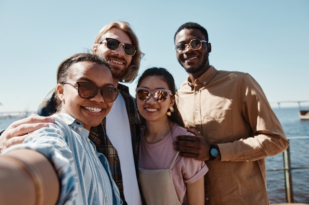 POV à un groupe diversifié de jeunes prenant un selfie à l'extérieur en été, tous portant des lunettes de soleil